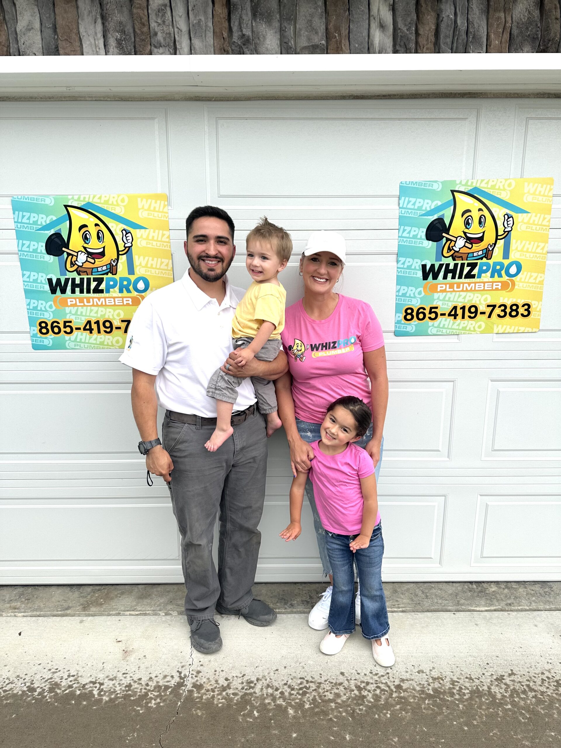 A man, woman, and two young children stand smiling in front of a garage door with two Whiz Pro Plumber signs displayed.