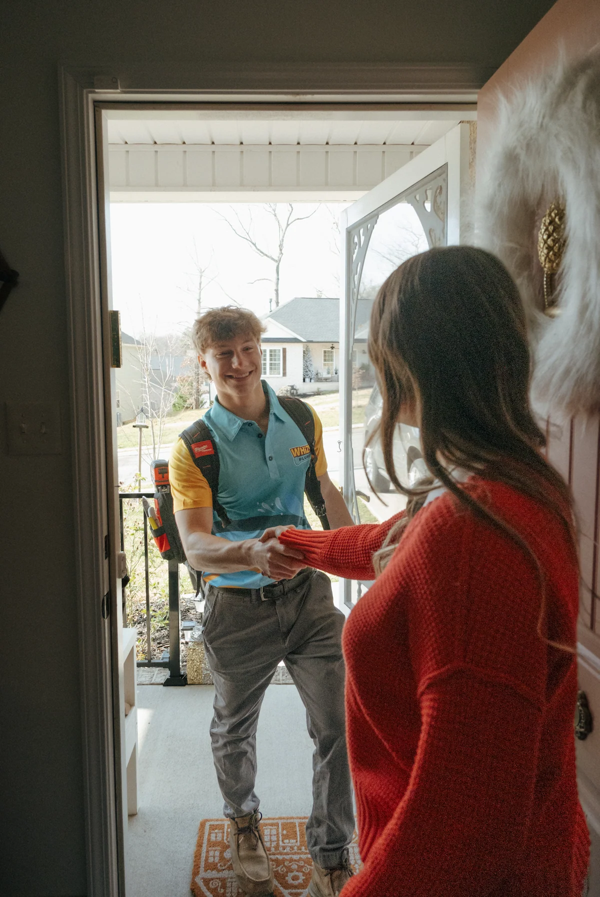 A young man with a backpack stands at the front door, shaking hands with a woman in a red sweater who is greeting him inside the house.