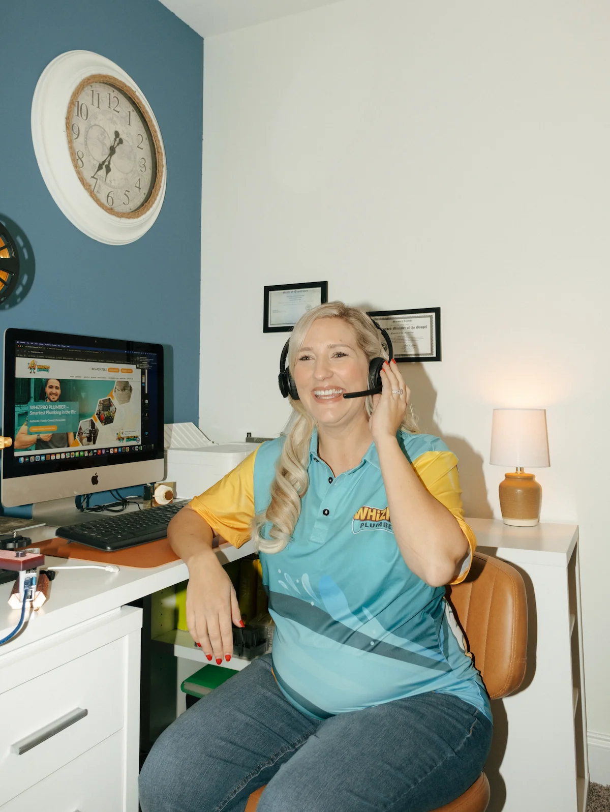 A woman wearing a headset sits at a desk in a home office, smiling at the camera. A computer, lamp, and wall clock are visible in the background.