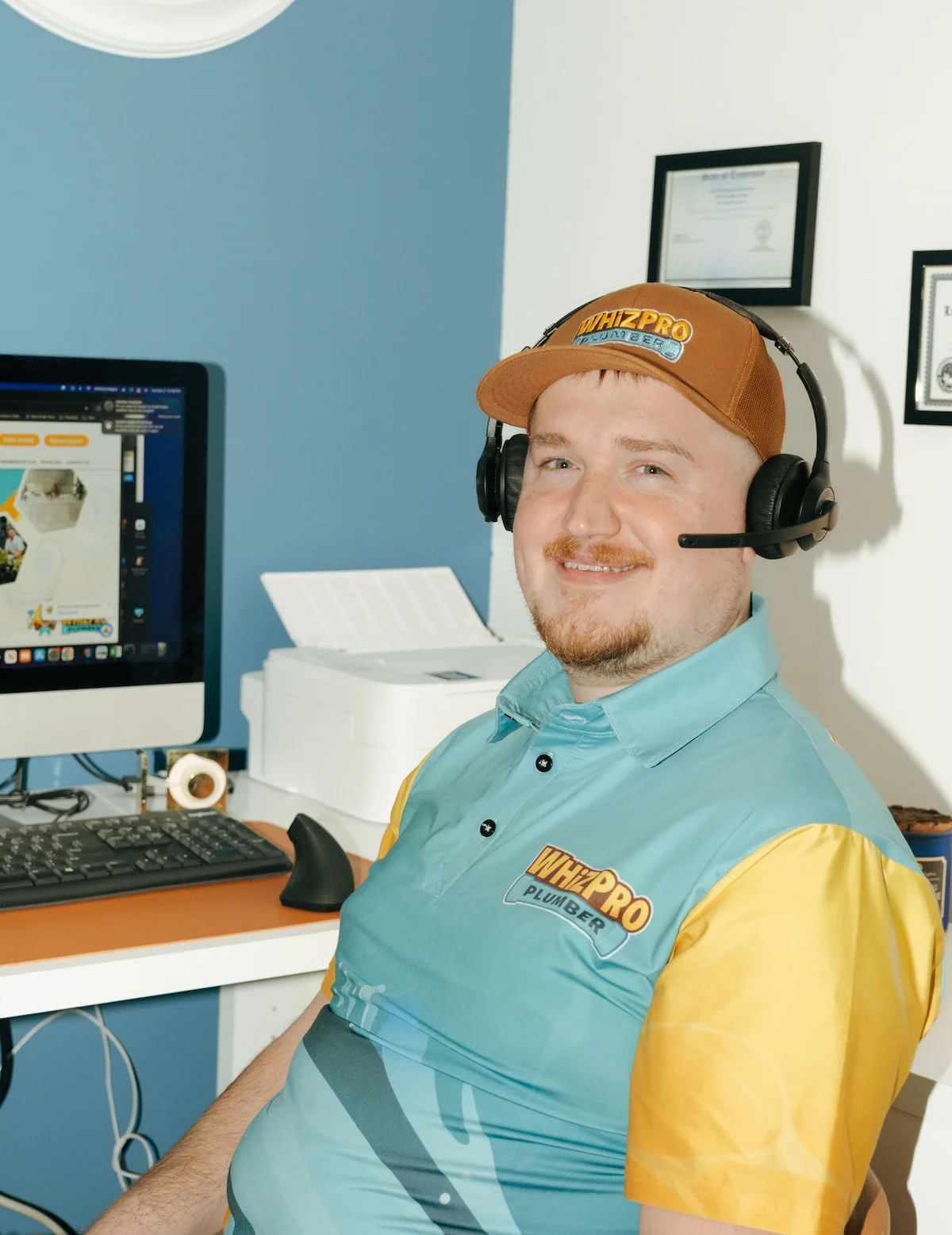 A man wearing a branded WHYZPRO polo shirt, cap, and headset sits at a desk with a computer and printer, smiling at the camera.