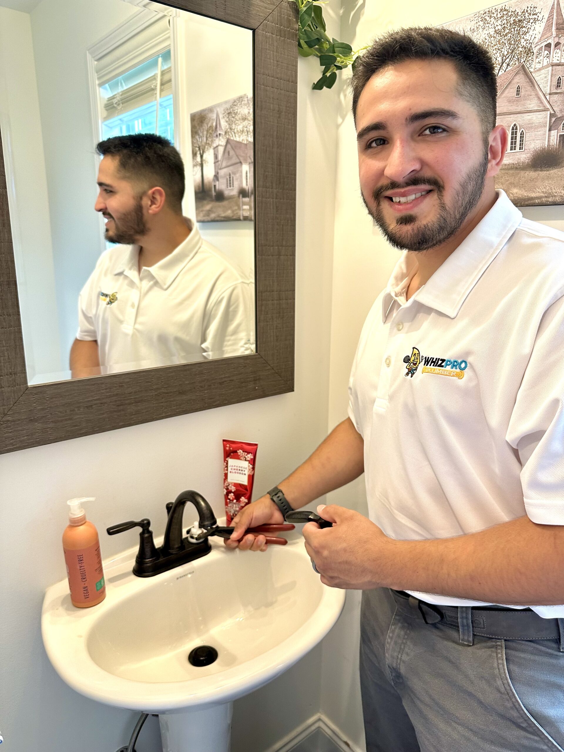 A man in a white polo shirt stands at a bathroom sink, using a wrench to fix the faucet while smiling at the camera.