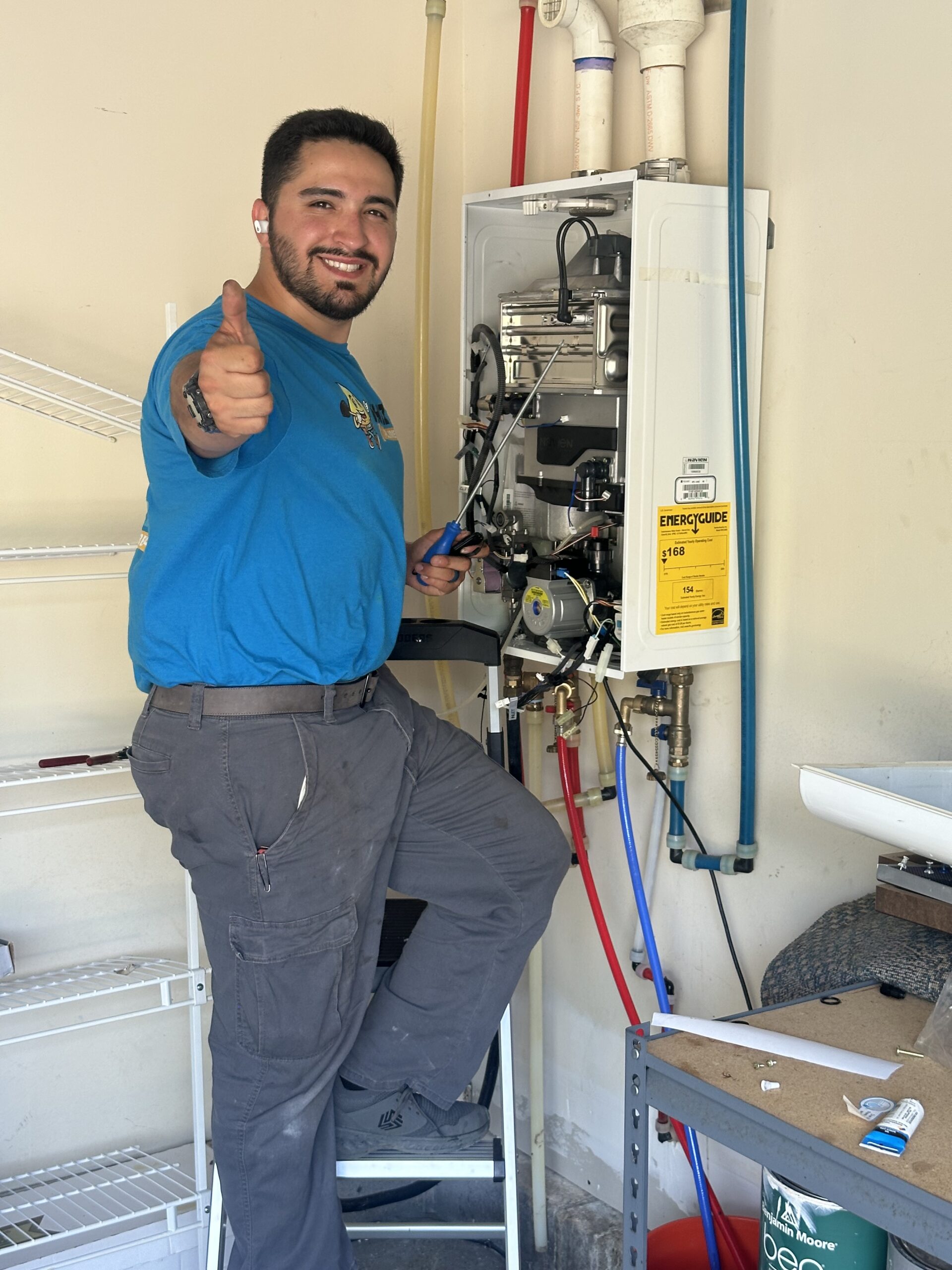 A technician in a blue shirt and gray pants stands on a step stool, smiling and giving a thumbs-up while working on a wall-mounted water heater in a garage.
