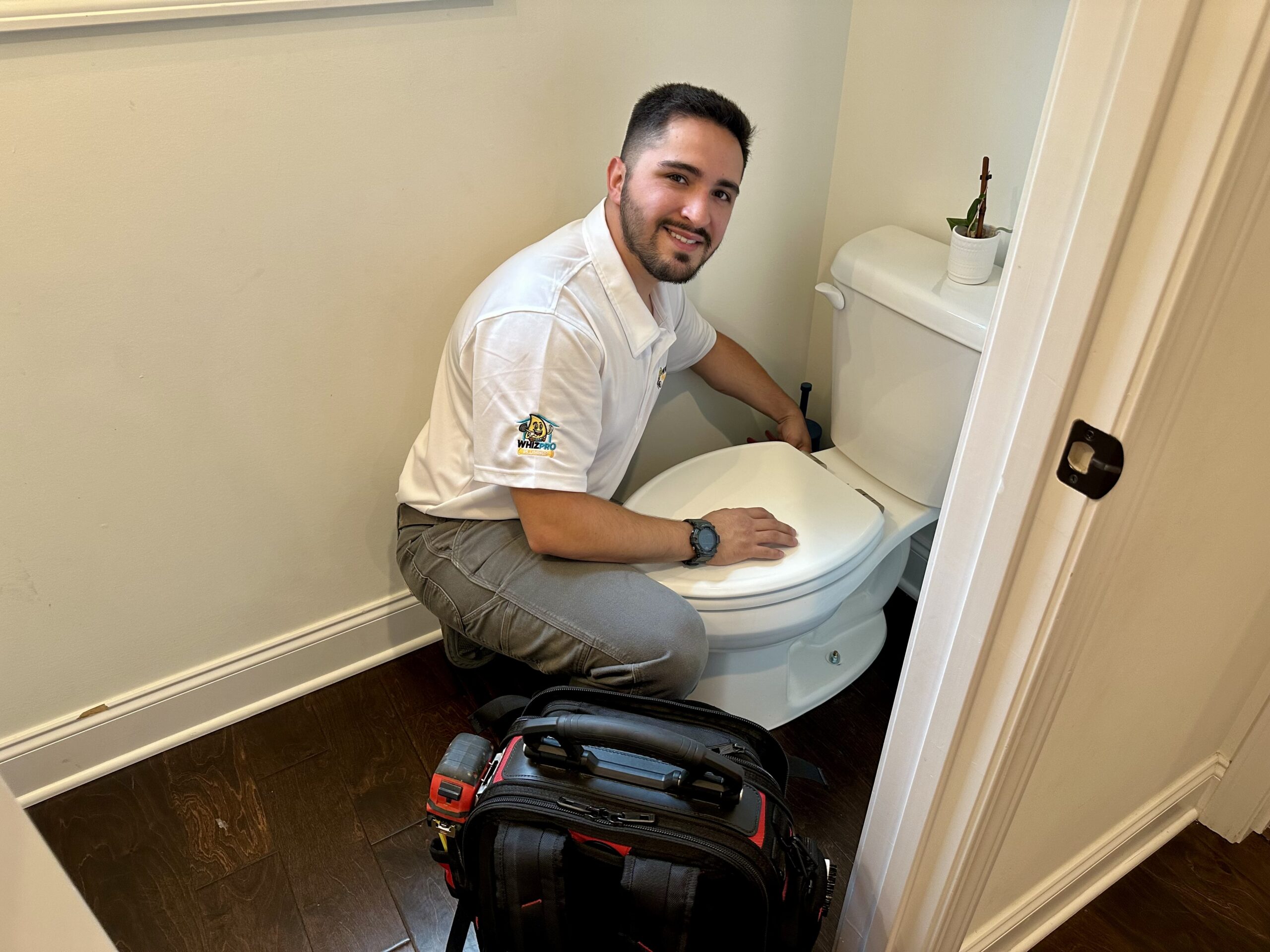 A man in a work uniform kneels next to a toilet, holding a tool, with a tool bag on the floor beside him in a small bathroom.
