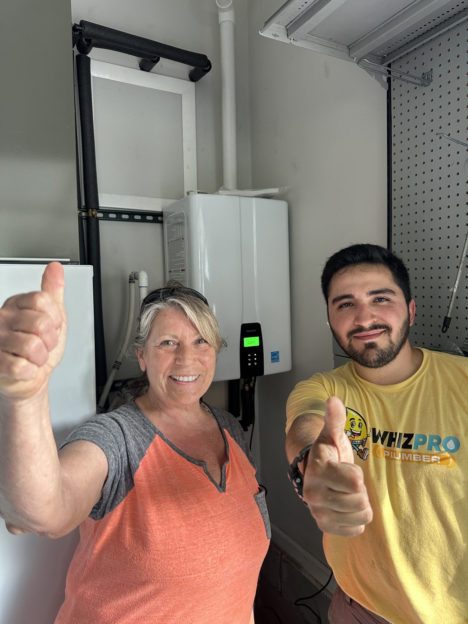 A woman and a man give thumbs up while standing in front of a wall-mounted appliance in a utility room.