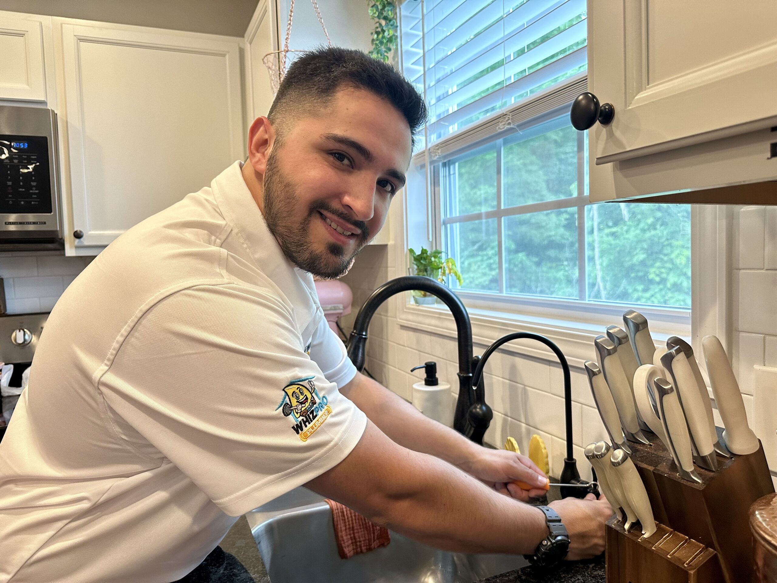 A man in a white shirt washes dishes at a kitchen sink, smiling at the camera. A knife block and window with greenery are visible nearby.