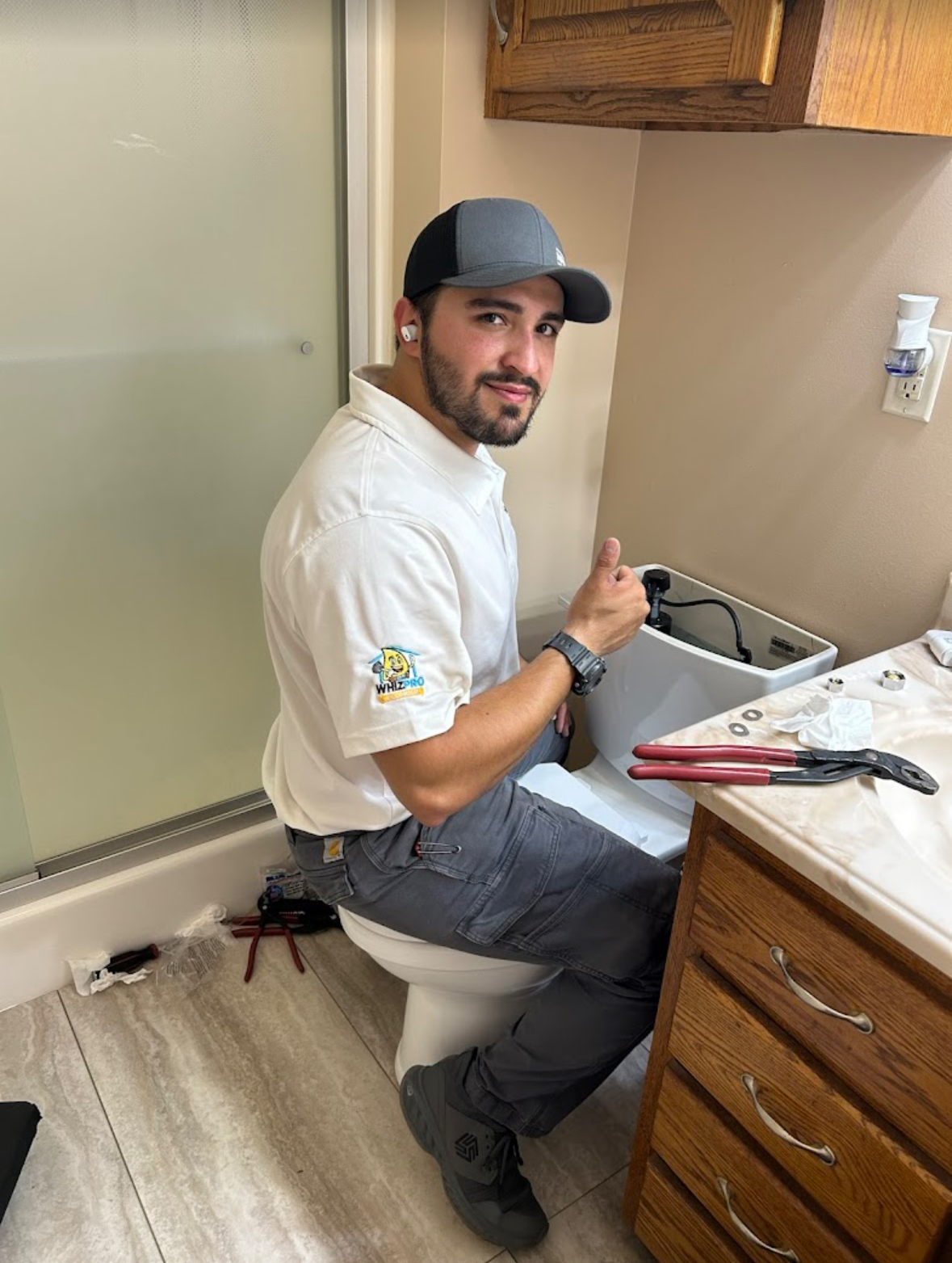A plumber in a white shirt and gray cap gives a thumbs-up while repairing a toilet in a bathroom; tools are placed on the floor and counter nearby.