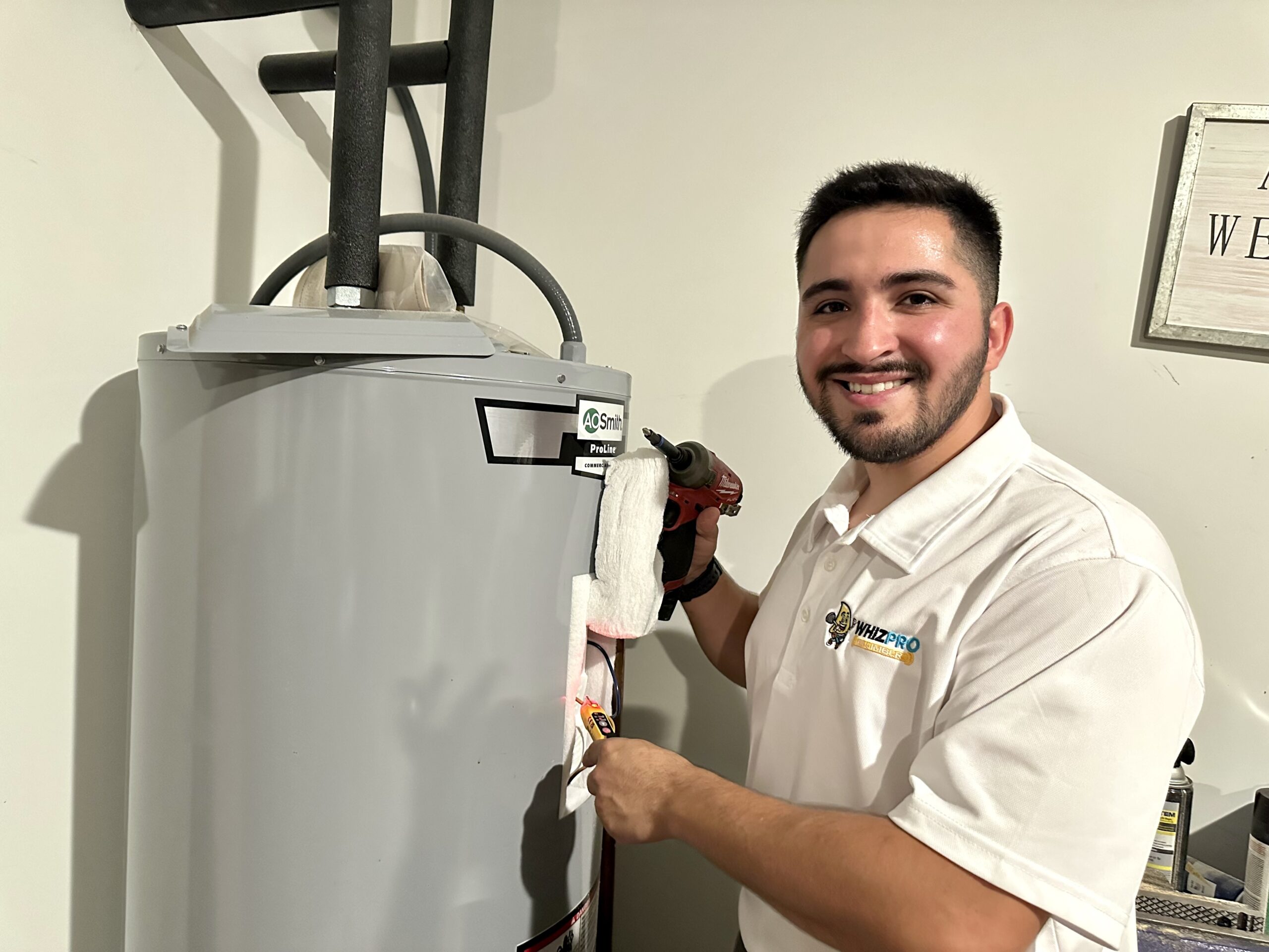 A man in a white polo shirt uses a tool to work on a water heater, smiling at the camera in a utility room.