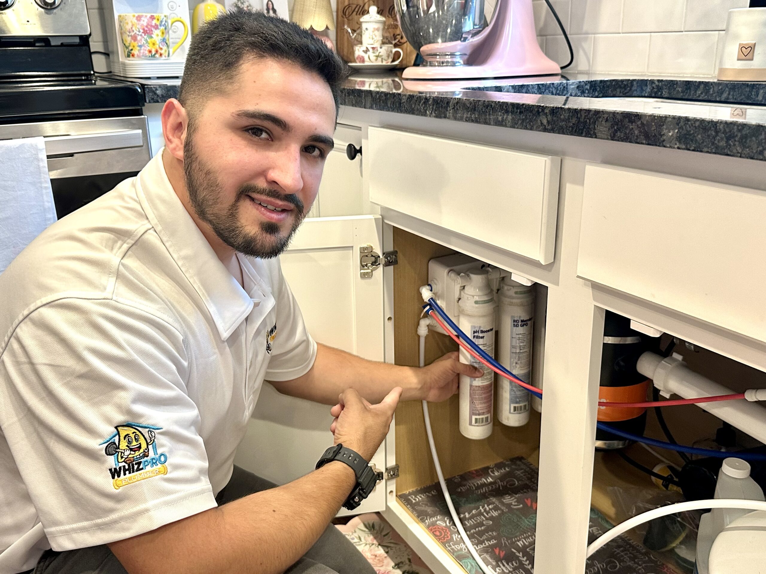 A technician kneels in a kitchen, pointing to a water filtration system installed under the sink.