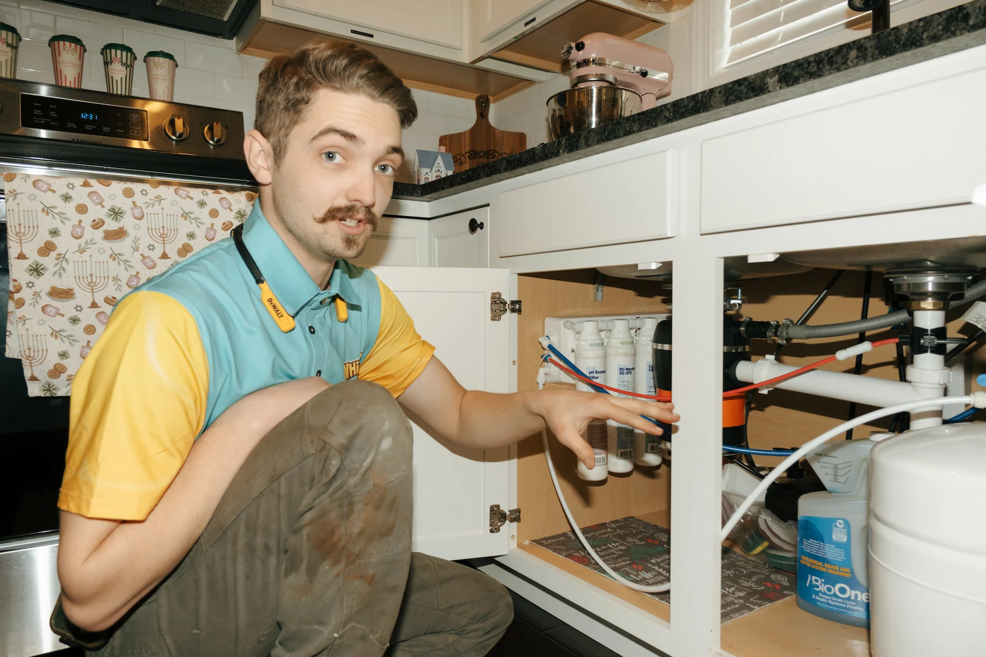 A man kneels in a kitchen, pointing to a water treatment system installed under the sink.