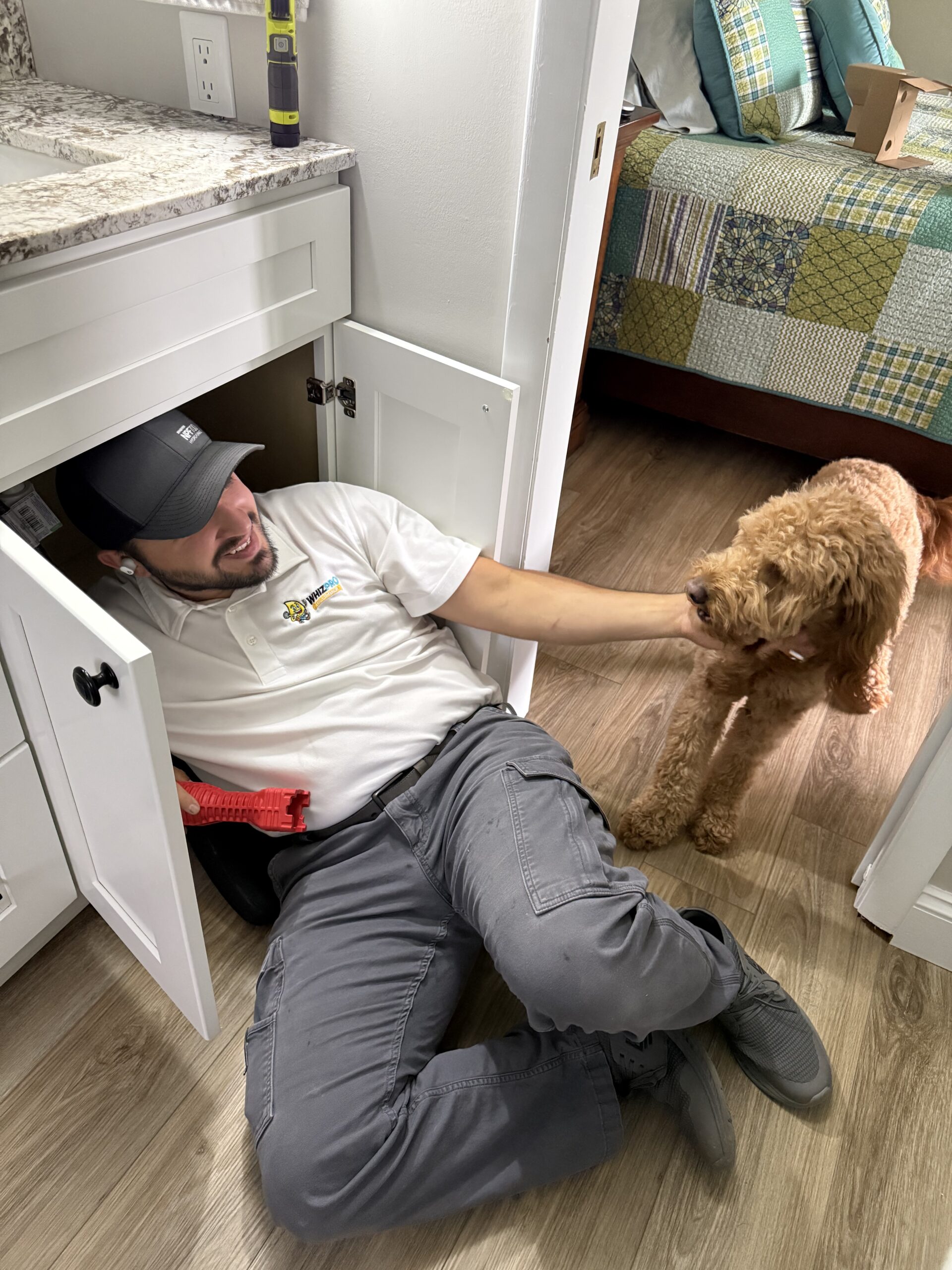 A man in work clothes lies under a sink cabinet, reaching out to pet a brown dog in a bedroom with a patterned bedspread.