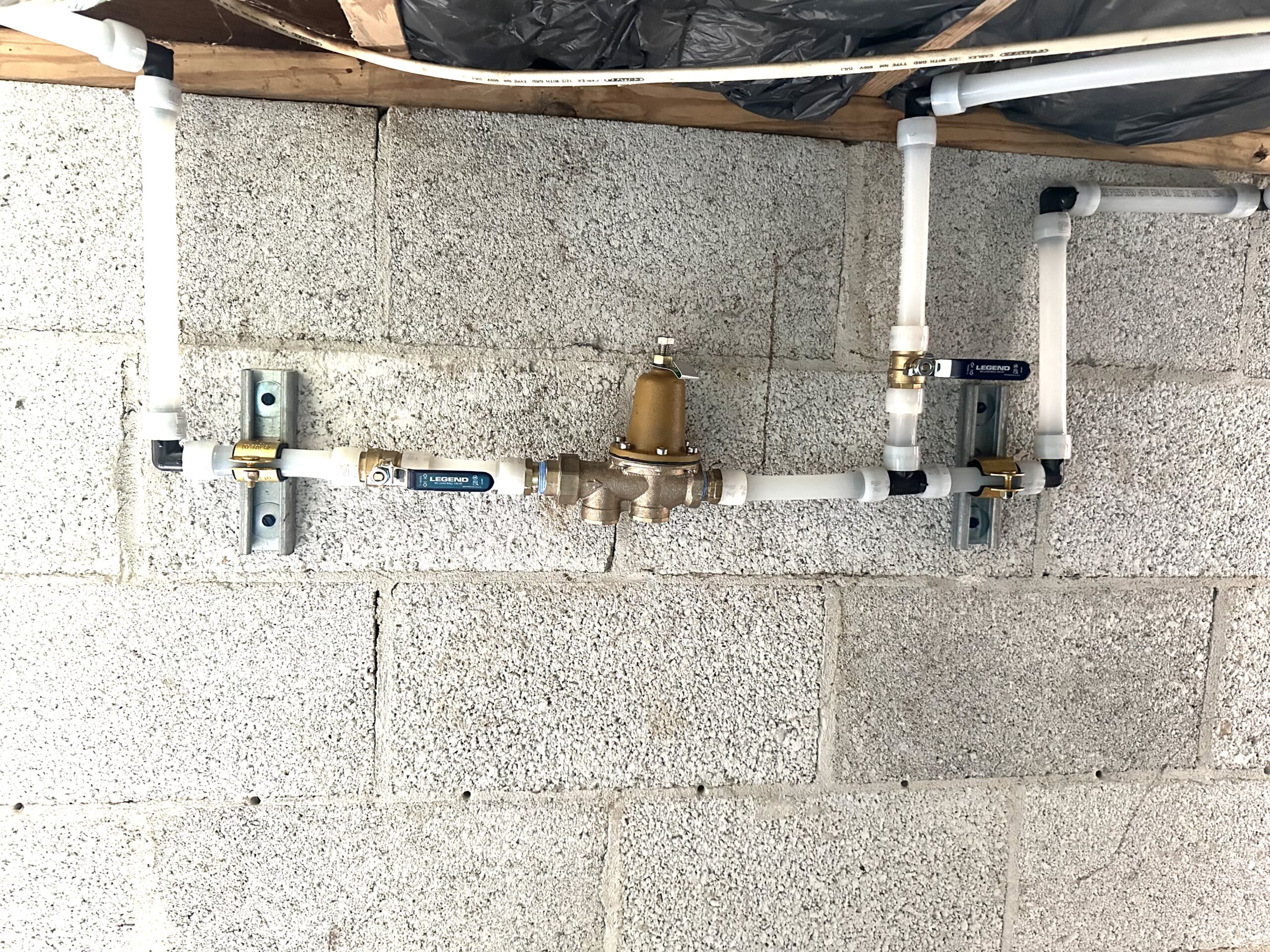 A water pressure regulator with connected PVC pipes and shut-off valves is mounted on a cinder block wall in a basement or utility room.